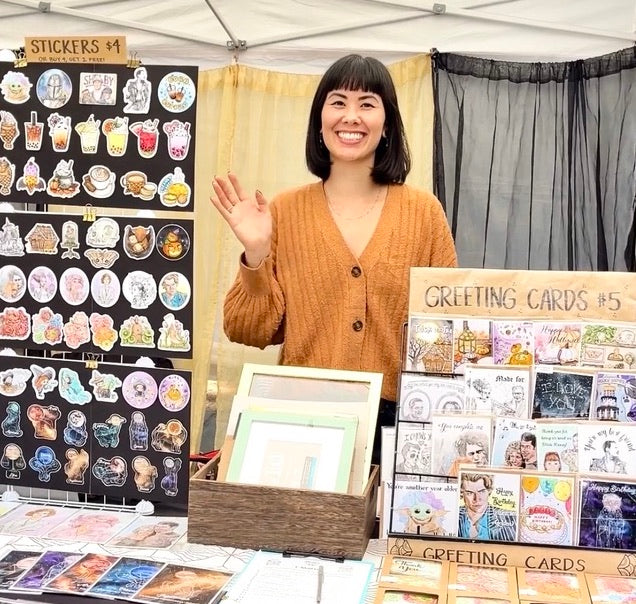 Photo of the artist, Monique Tiffer, smiling and waving from her art booth at a makers market.