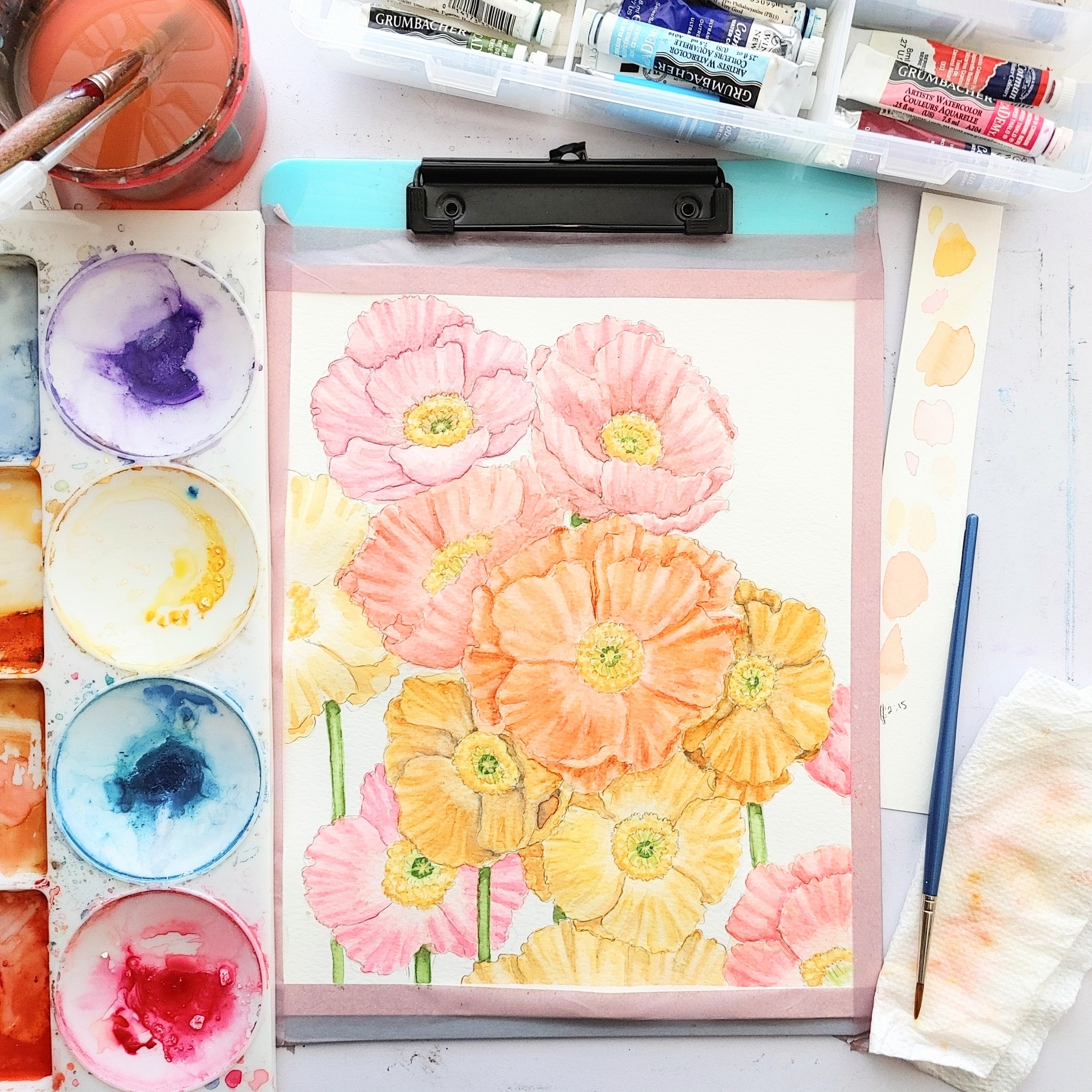 Process shot of colorful pink, orange, and yellow poppies being painted with watercolor. Watercolor paints and brushes sit on the table.