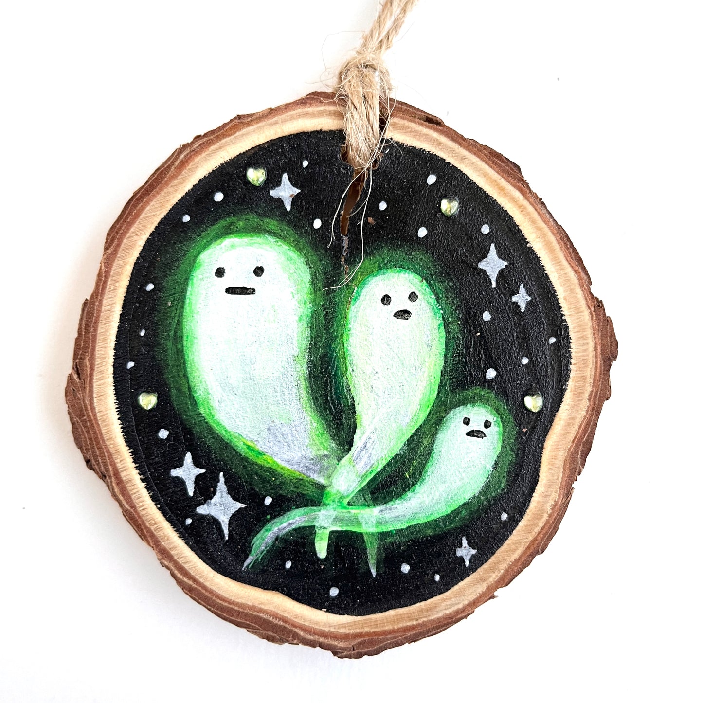 Three ghost that appear to be glowing decorate this wooden ornament with green heart rhinestones on a black background.