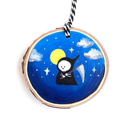 Wooden ornament with a painted cute small grim reaper with his scythe. A full moon and stars and clouds are in the dark blue background.