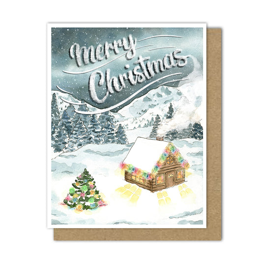 A Christmas card of a wooden cabin in the snow with Christmas lights, and snowy tree outside with Christmas lights. A galaxy hangs aboce the snowy mountains with Merry Christmas written in the cabin's smoke coming out of the chimney.