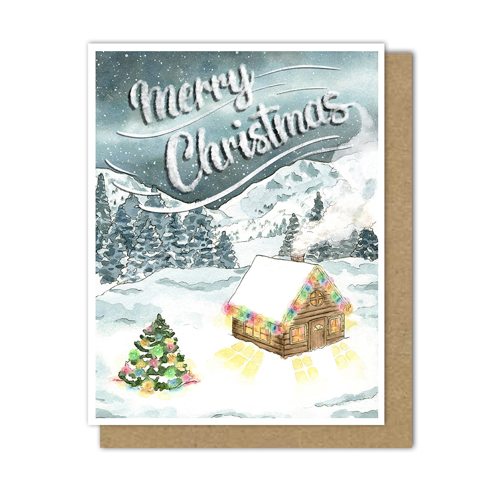 This festive card showcases a star studded galaxy floating above a quaint cabin in the snow. Colorful rainbow lights adorn the cabin rooftop and a nearby tree from the snowy forest. 