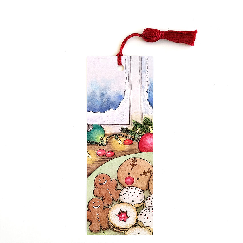 Red tassel bookmark featuring a plate of Christmas cookies on a table with ornaments and lights in front of a snowy window.