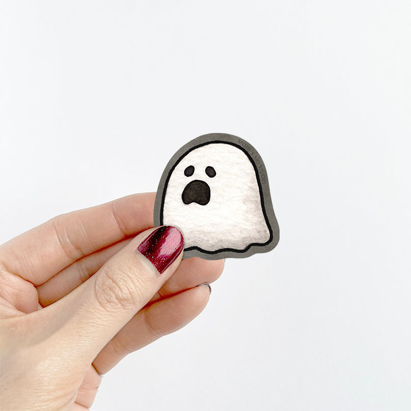 A hand holding a small sticker of a white moaning ghost that has a dark gray border.