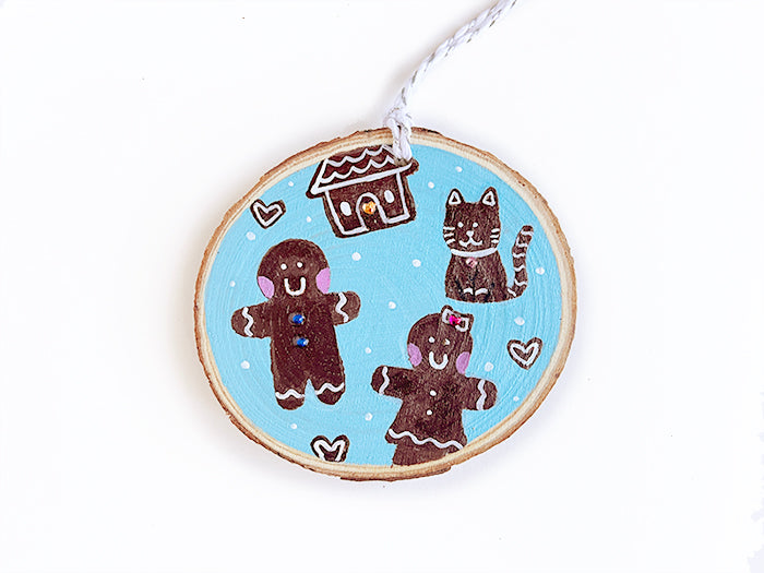 Wooden ornament with hand painted gingerbread man, woman, house, cat, and hearts on a blue background.