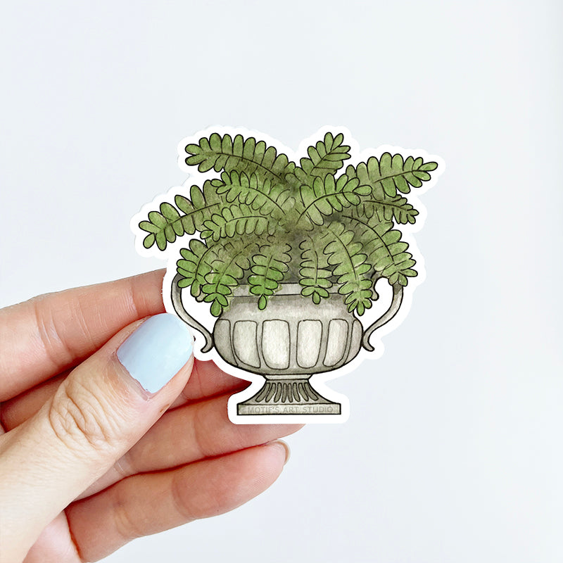 Hand holding a sticker of a Boston fern inside a gray stone urn.