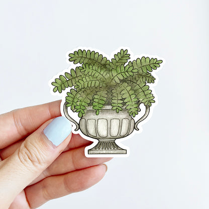 Hand holding a sticker of a Boston fern inside a gray stone urn.