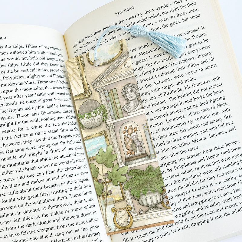 Bookmark with blue tassel featuring a greek mythology inspired bookcase, art, and plants. The bookmark is placed inside a book.