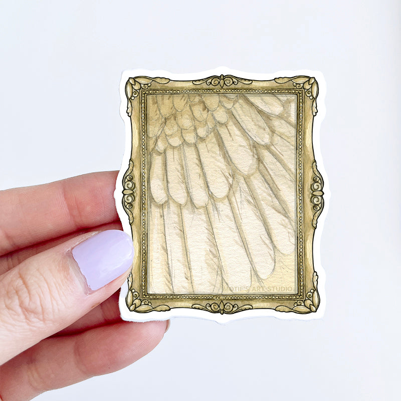 Hand holding a sticker of a vintage frame of a golden wing.