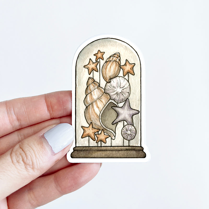 Hand holding a sticker of a variety of orange seashells, sand dollars, and starfish inside a terrarium.