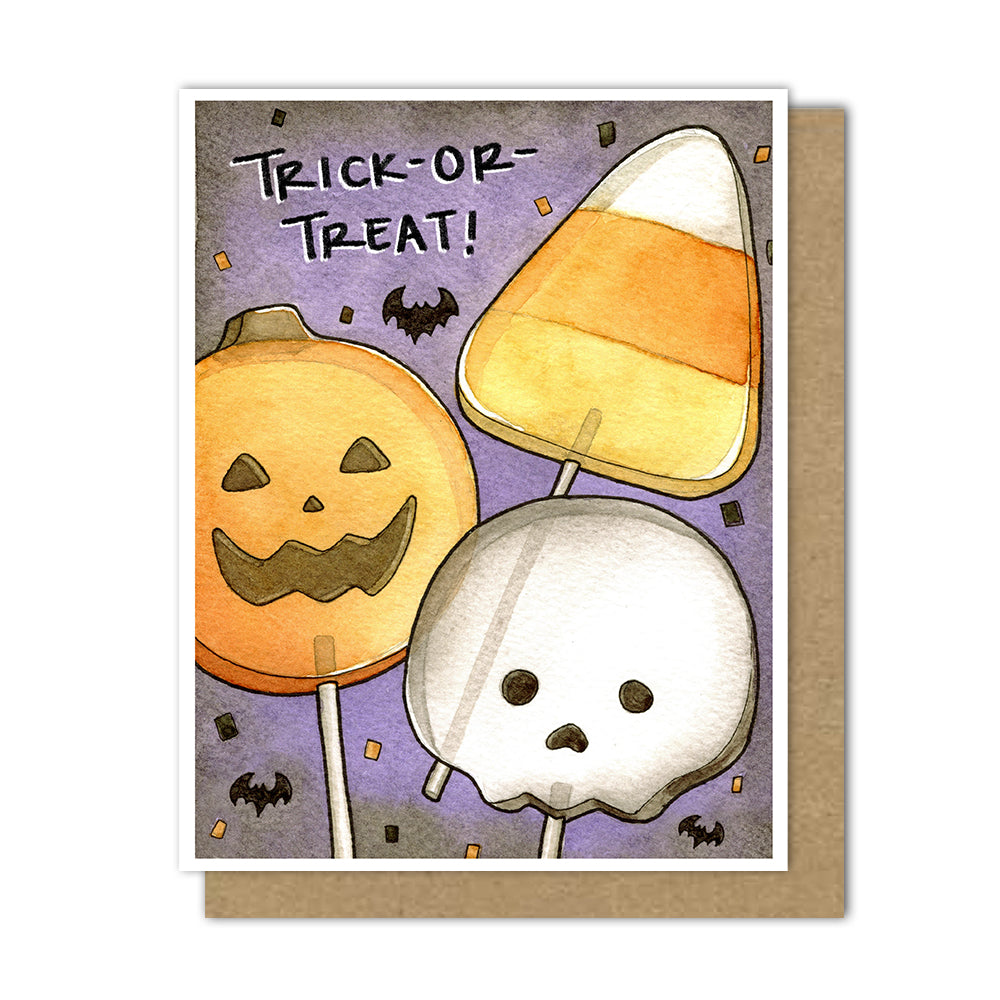 An A2 Halloween greeting card with a white border, featuring watercolor-style illustrations of a jack-o'-lantern lollipop, a candy corn lollipop, and a skull lollipop on a purple background with the words “TRICK-OR-TREAT!” A kraft brown envelope is placed behind the card.