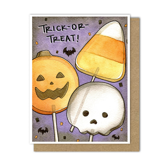 An A2 Halloween greeting card with a white border, featuring watercolor-style illustrations of a jack-o'-lantern lollipop, a candy corn lollipop, and a skull lollipop on a purple background with the words “TRICK-OR-TREAT!” A kraft brown envelope is placed behind the card.