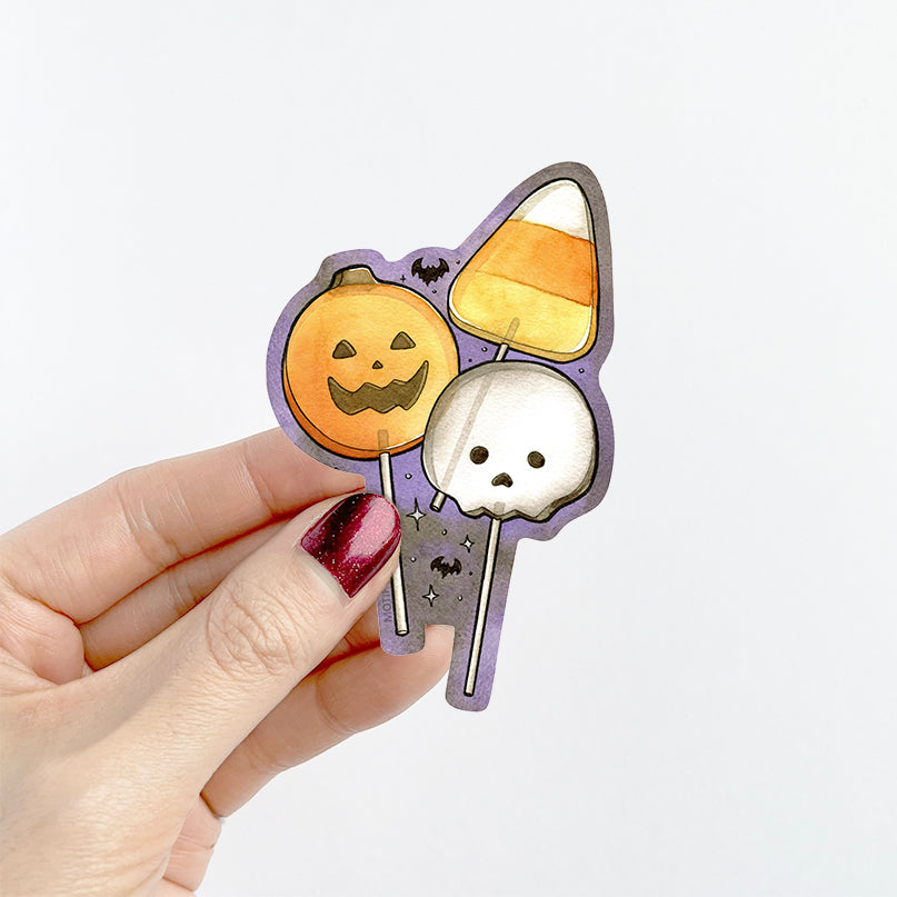 A hand with dark glittery nail polish holds a die-cut sticker featuring a cute Halloween illustration. The sticker shows a smiling orange pumpkin, a candy corn, a white skull, and little black bats on a purple background.