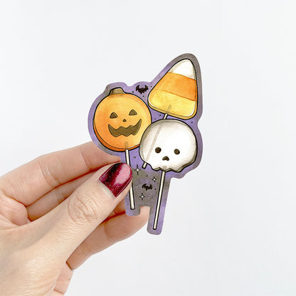 A hand with dark glittery nail polish holds a die-cut sticker featuring a cute Halloween illustration. The sticker shows a smiling orange pumpkin, a candy corn, a white skull, and little black bats on a purple background.