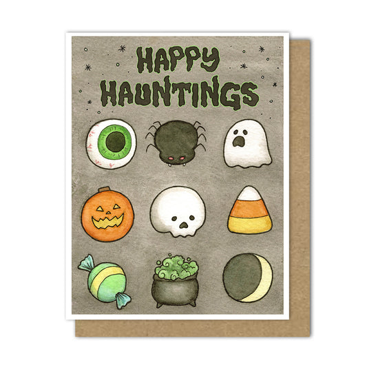 A Halloween greeting card with a brown kraft envelope behind it. The card reads “Happy Hauntings” in dark green textured lettering above a grid of cute illustrated spooky treats, including a green-and-white eyeball, a black spider, a white ghost, an orange jack-o'-lantern, a white skull, candy corn, a striped candy, a cauldron of green potion, and a crescent moon, all on a gray background.