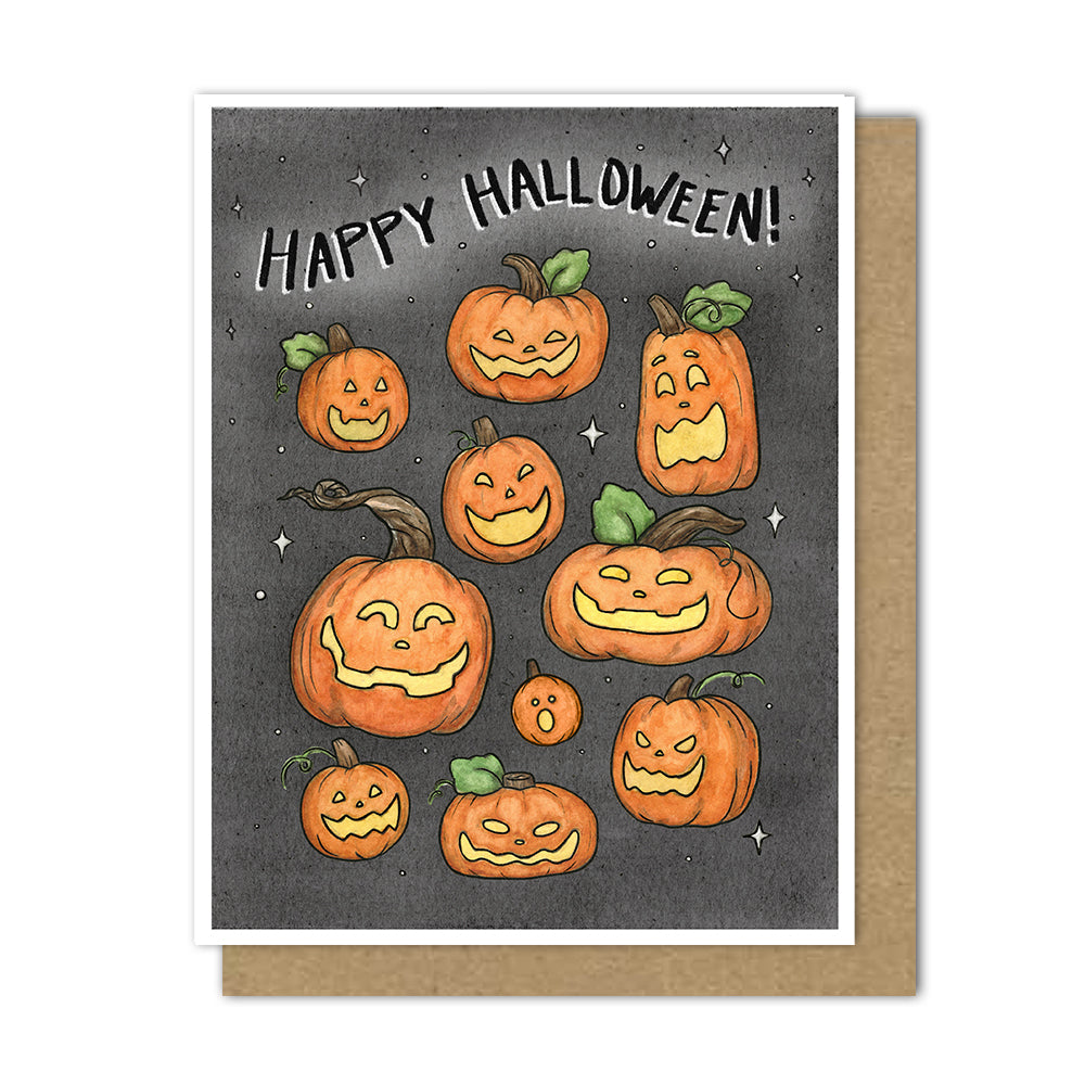 A Halloween greeting card with a white border, featuring illustrated jack-o'-lantern pumpkins on a dark gray background with the words 'Happy Halloween!' at the top, shown with a brown kraft envelope behind it.