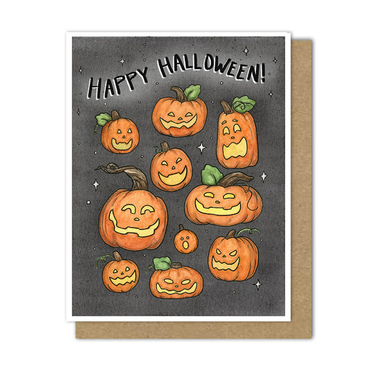 A Halloween greeting card with a white border, featuring illustrated jack-o'-lantern pumpkins on a dark gray background with the words 'Happy Halloween!' at the top, shown with a brown kraft envelope behind it.