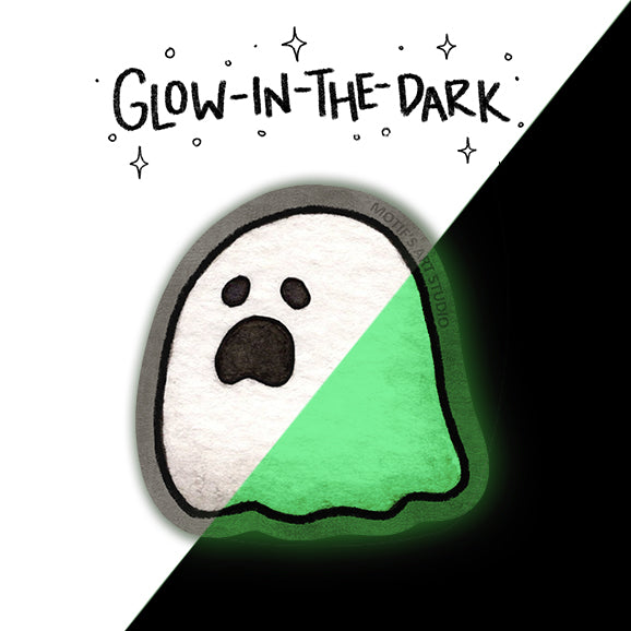 A ghost-shaped glow-in-the-dark sticker shown on a split white-and-black background with the text 'GLOW-IN-THE-DARK' above it. The ghost is white on the left and glows green on the right, with a dark mouth and simple black facial features.