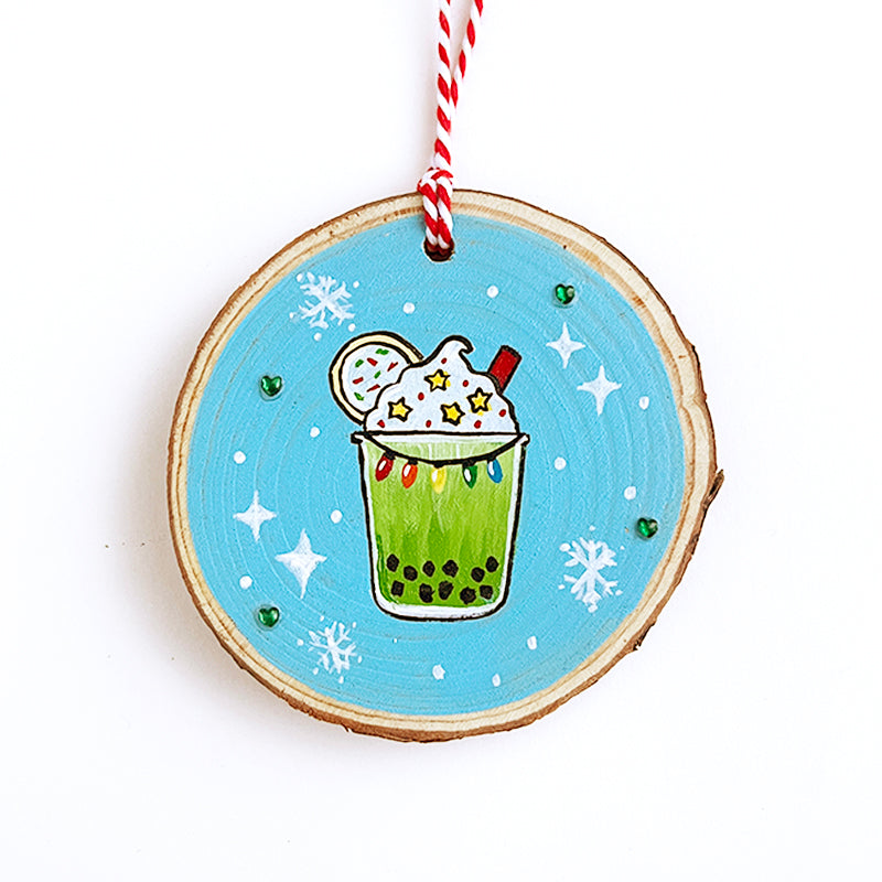 Wooden ornament with matcha green boba drink with stars in the whipped cream and lights hanging on the drink. The background has snowflakes and green rhinestones.