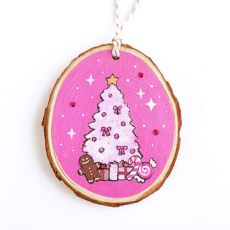 Wooden ornament with painted light pink Christmas tree, medium pink background and sparkles and magenta rhinestones. The tree has a gingerbread man, presents, and peppermints under the tree.