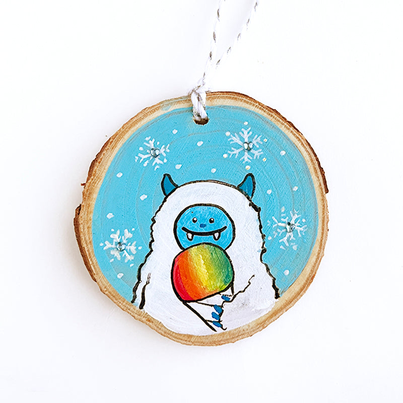 Wooden round ornament with a blue and white smiling yeti eating a rainbow snowcone with snowflakes in the blue background.