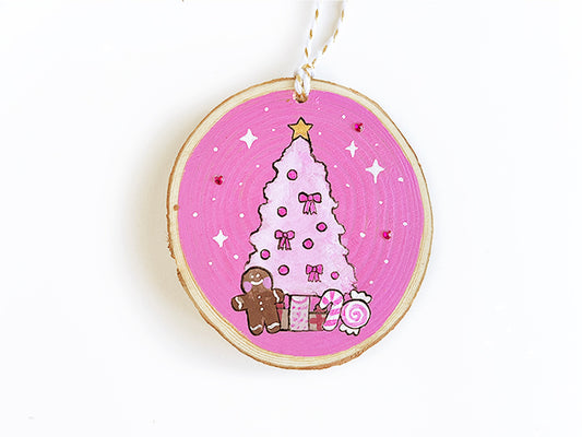 Wooden ornament with painted light pink Christmas tree, medium pink background and sparkles and magenta rhinestones. The tree has a gingerbread man, presents, and peppermints under the tree.