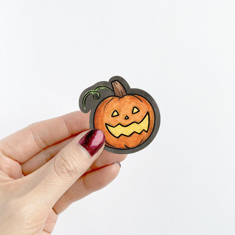 A hand with glittery dark red nail polish holds a small die-cut sticker of a hand-drawn orange jack-o'-lantern pumpkin with a black outline against a plain white background.