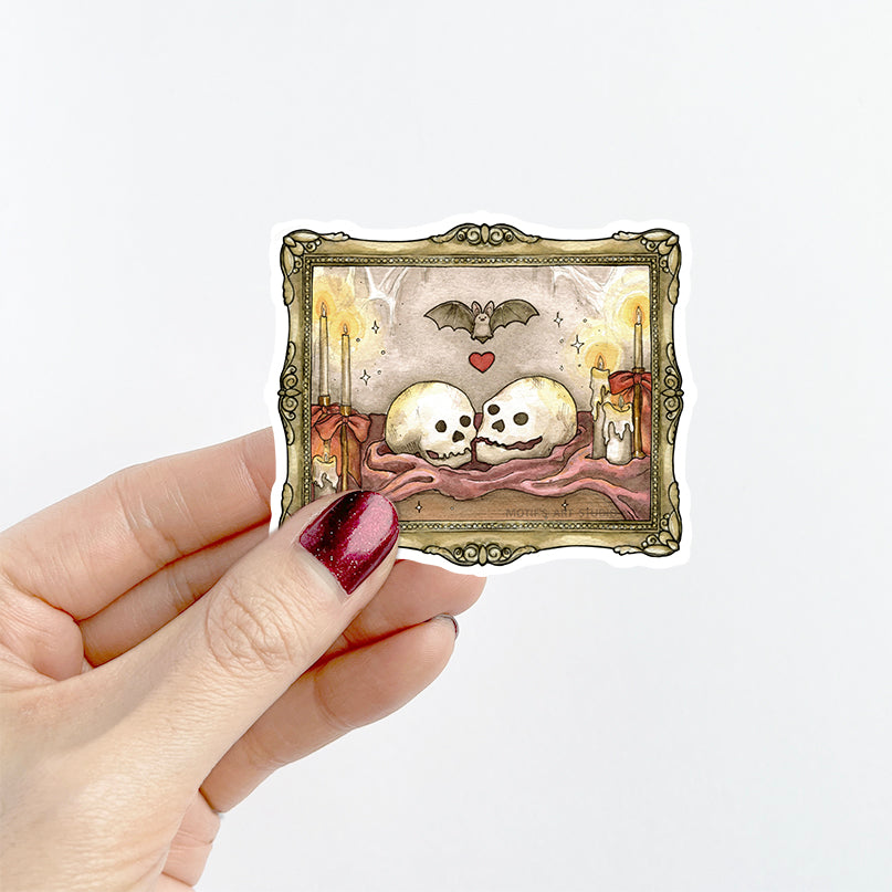A hand with dark red glitter nail polish holds a die-cut sticker featuring a gothic illustration of two skulls, candles, a bat, and a small red heart inside an ornate frame, against a plain white background.