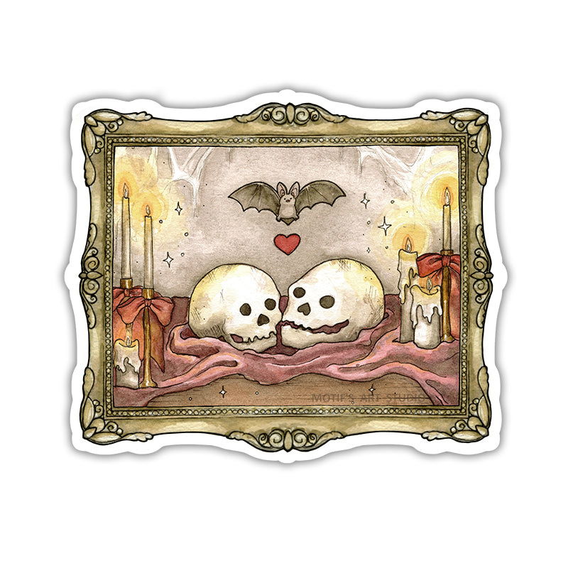 A die-cut sticker featuring a gothic illustration of two skulls resting on a maroon cloth, flanked by lit candles, with a bat and a red heart above, all framed by an ornate gold frame.