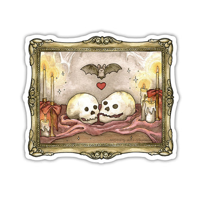 A die-cut sticker featuring a gothic illustration of two skulls resting on a maroon cloth, flanked by lit candles, with a bat and a red heart above, all framed by an ornate gold frame.