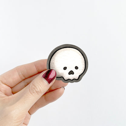 A hand holding a small sticker of a white skull with a dark gray border.