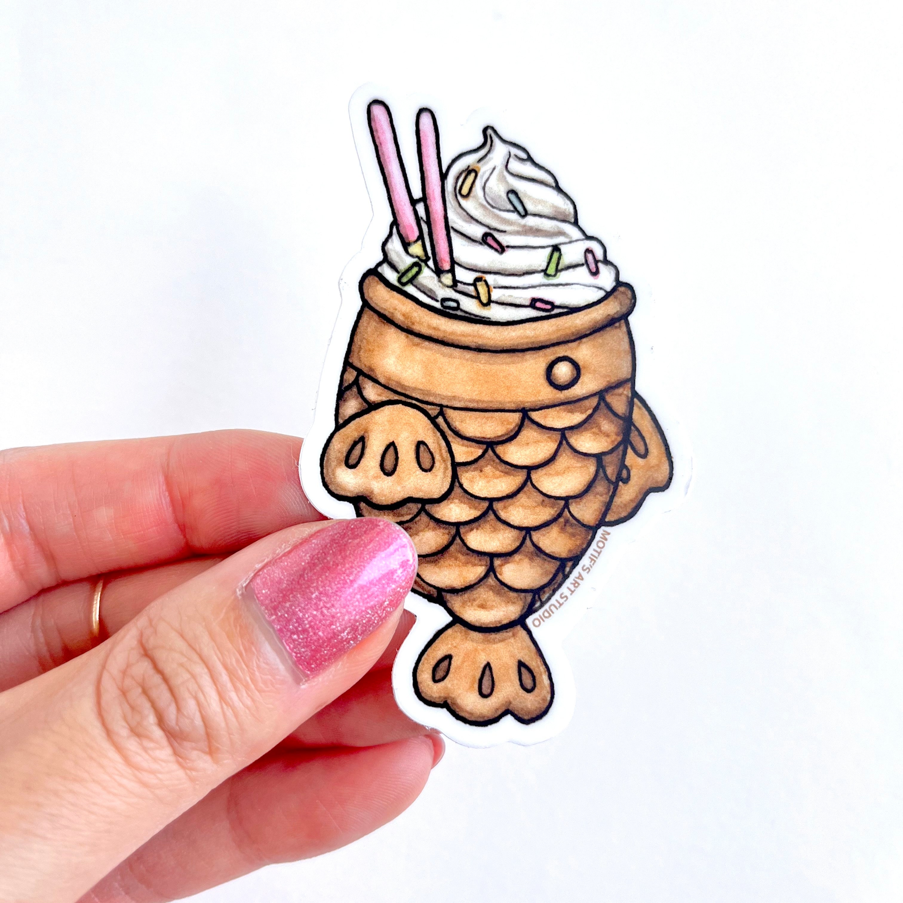 A hand holding up a vinyl sticker of a cute taiyaki fish cone with pink pocky stickers and sprinkles.