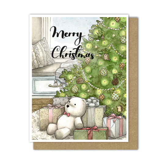 Greeting card with lush christmas tree with presents under the tree and a polar bear stuffed animal. A knit pillow and blanket sit in the background on a bench looking out a snowy window.