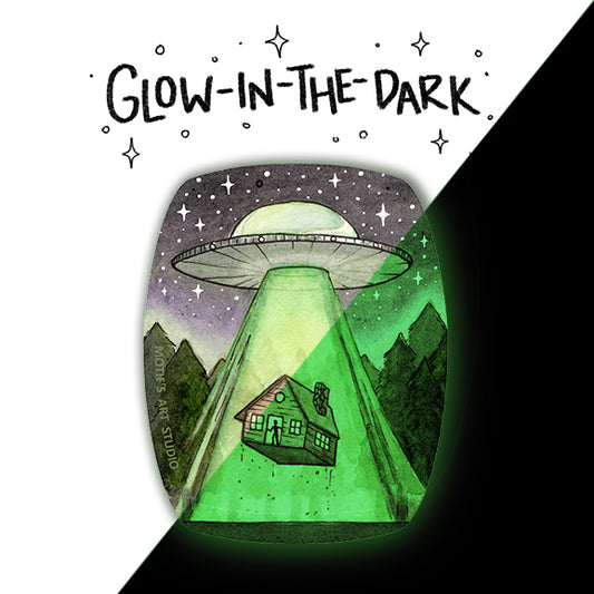 An oval shaped sticker of a glow in the dark UFO abducting a cabin in the forest.