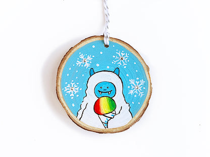 Wooden round ornament with a blue and white smiling yeti eating a rainbow snowcone with snowflakes in the blue background.